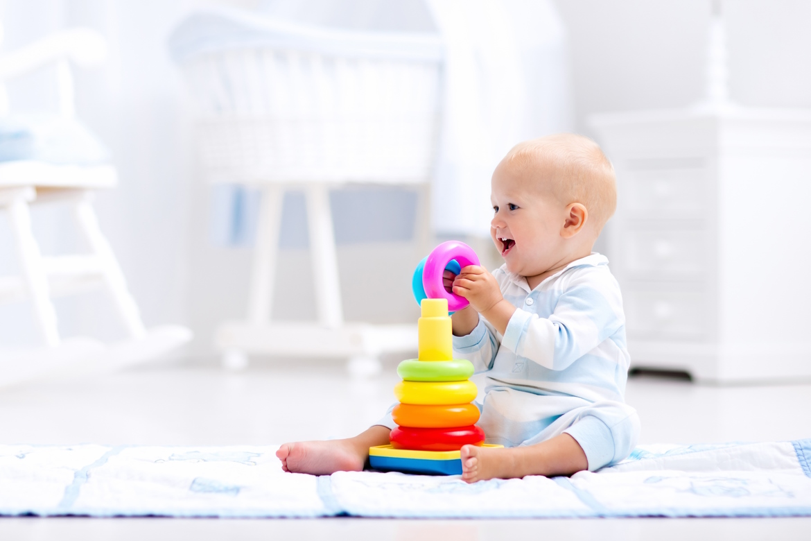 Das süße Baby spielt mit einer bunten Regenbogen-Spielzeugpyramide und sitzt auf Spielmatte im weißen, sonnigen Schlafzimmer. Spielzeug für kleine Kinder. Inneneinrichtung für kleine Kindergarten.